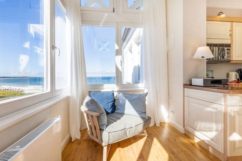 A room with a view of the sea, a wooden chair with cushions, a kitchen area with cabinets, and a radiator.