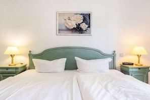 Ein ordentlich bezogenes Bett mit weißen Leinen, zwei Nachttischlampen und ein gerahmtes Blumenbild an der Wand.