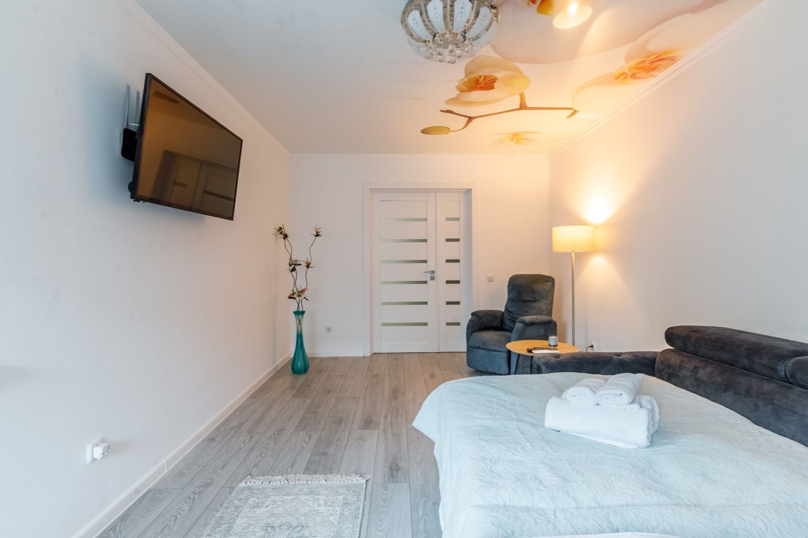 Une chambre d’hôtel comprenant un lit, un coin salon avec une chaise et une table, un vase avec des fleurs, un téléviseur à écran plat fixé au mur et une porte à motifs rayés.