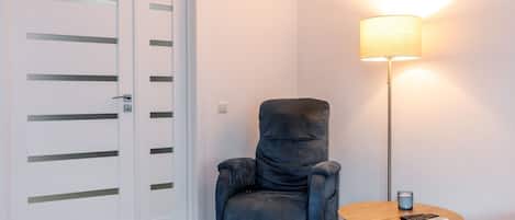 Una stanza con una poltrona blu, un tavolino rotondo con una lampada e una porta bianca con listelli orizzontali.