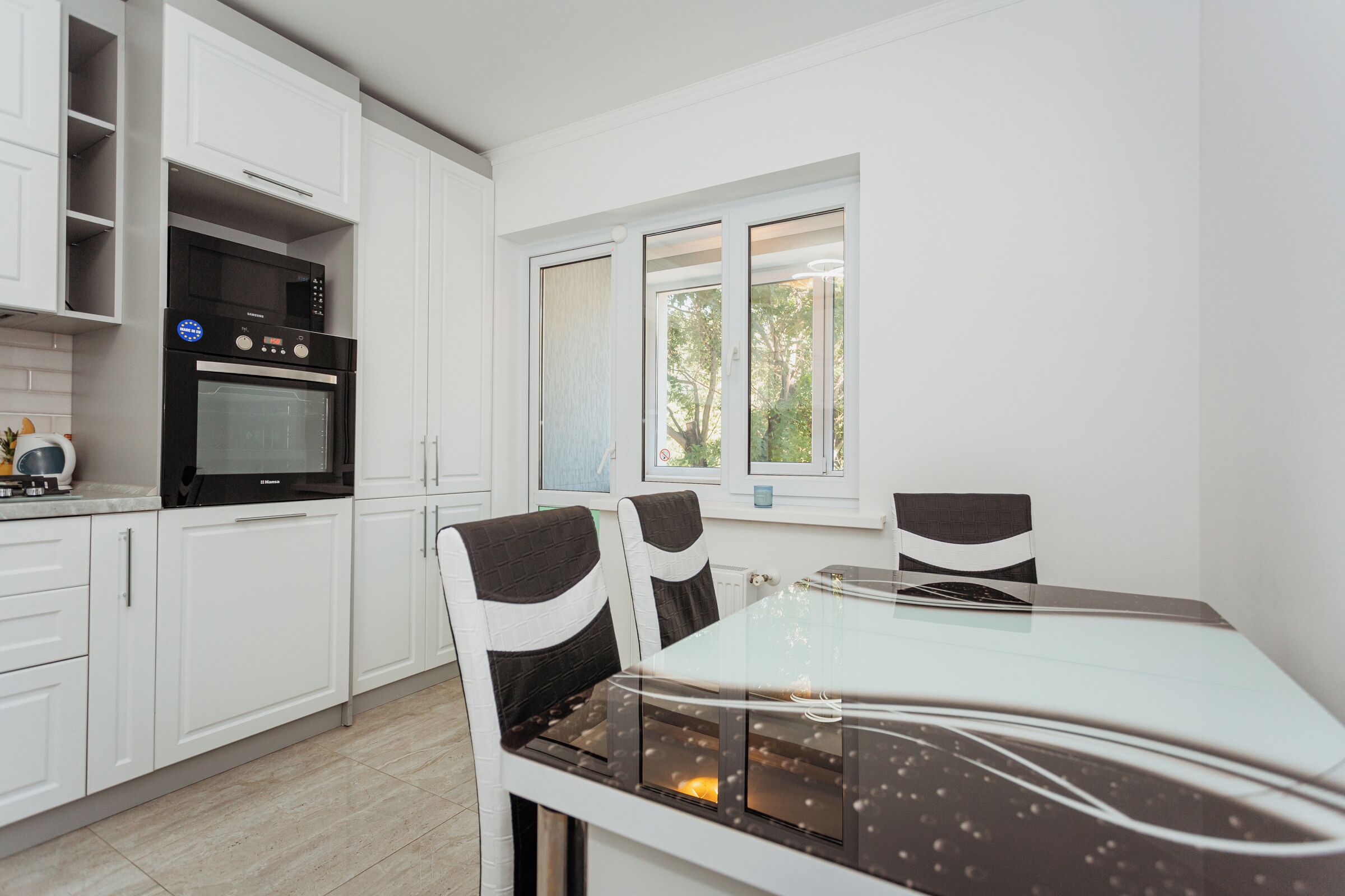 Een moderne keuken met witte kasten, een inbouwoven en een eethoek met een glazen tafel en stoelen.