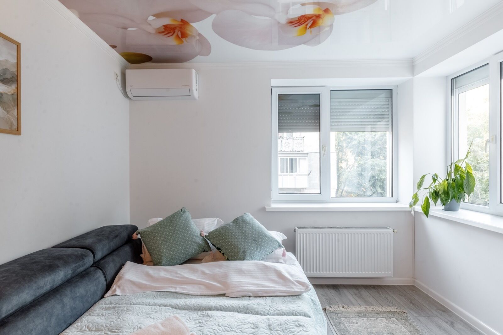 Een slaapkamer met een bed, een radiator, een raam met zonwering en een plant op het raamkozijn.