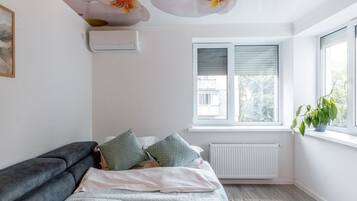 Een slaapkamer met een bed, een radiator, een raam met zonwering en een plant op het raamkozijn.