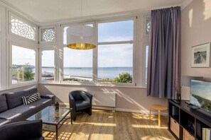 A modern living room with a sofa, a chair, a glass coffee table, and a television. There are large windows with a view of the sea and a bridge. - Stella Maris, Villa Agnes (Binz)