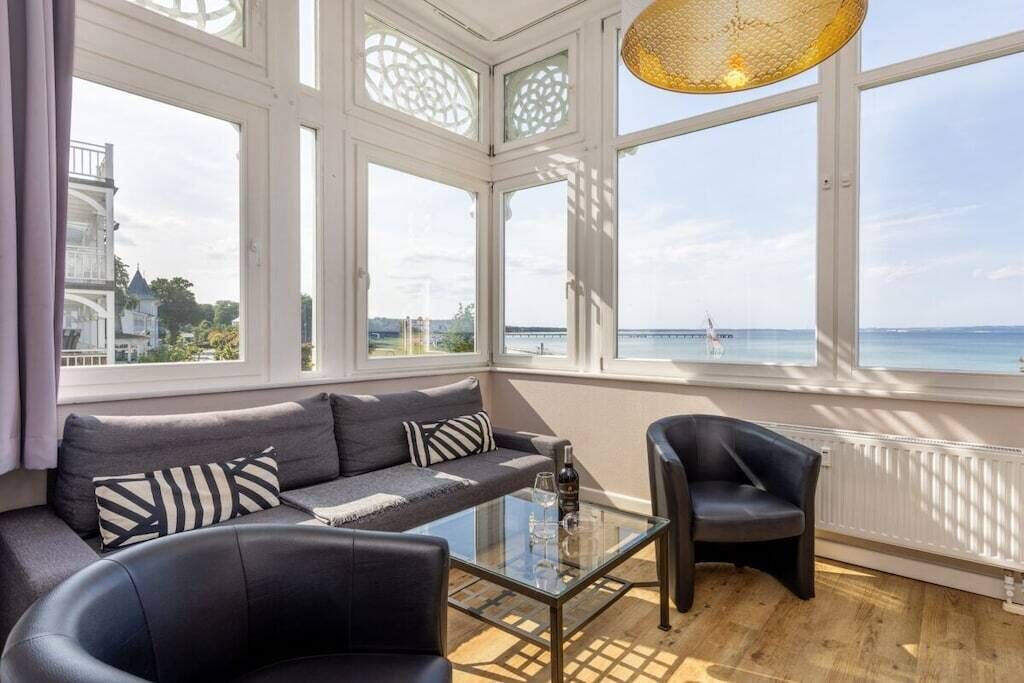 Ein modernes Wohnzimmer mit einer Couch, einem Glastisch und einem Sessel, große Fenster mit Blick auf das Meer und ein Schiff in der Ferne.