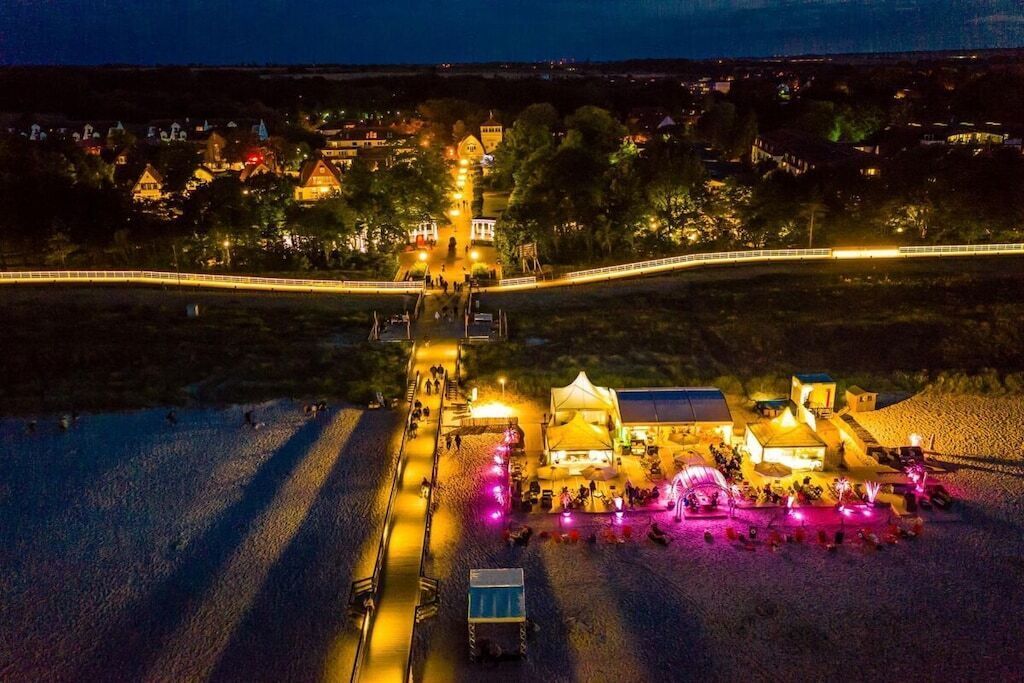 A nighttime view of a coastal area with a lit-up building complex, a bridge, and a beach with people.