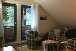 Ein gemütlicher Wohnbereich mit einer Couch, Sesseln und einem kleinen Tisch. Eine Tür führt zu einem Balkon mit Blick auf Grünflächen.