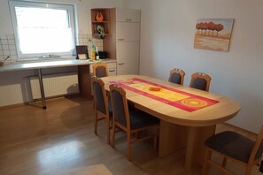 Een eetruimte met een ronde tafel, zes stoelen, een raam, een radiator, een kast en een schilderij aan de muur.
