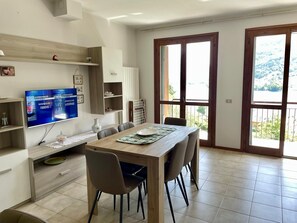 Un coin repas avec une table en bois, des chaises, un téléviseur fixé au mur et une vue sur l’extérieur grâce à de grandes fenêtres.