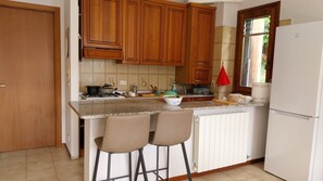 Une cuisine avec des meubles de rangement en bois, un réfrigérateur blanc, un plan de travail avec un saladier, et deux tabourets de bar.