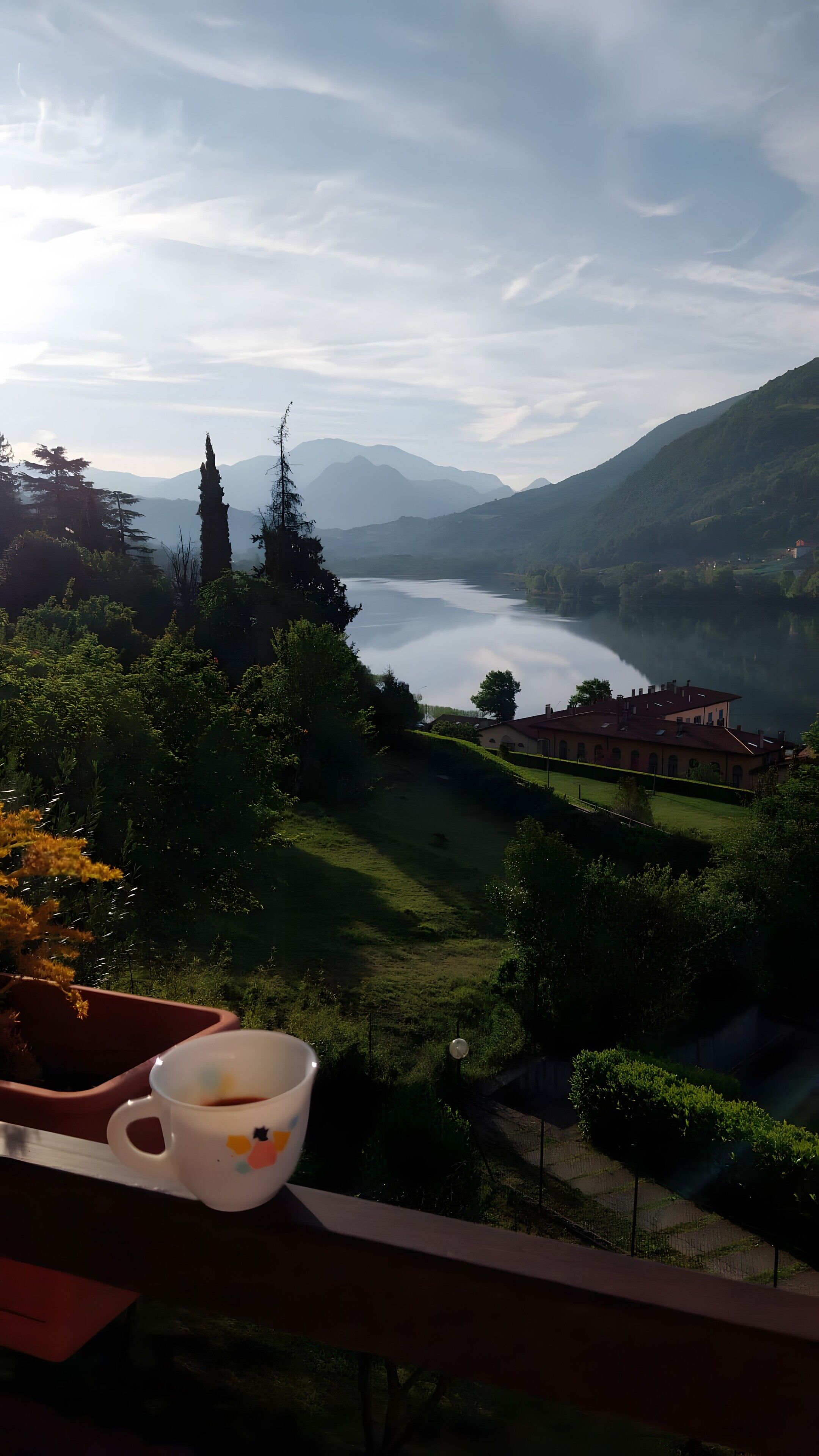 Ein malerischer Blick auf einen See mit Bergen im Hintergrund, eine Tasse auf einem Balkon und ein Gebäude mit rotem Dach.
