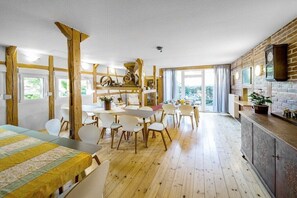 A spacious room with a wooden floor, a dining area with white chairs, a kitchen area with a brick wall, and a large window providing natural light. - Holiday home with 1 bedroom (Königsee \/ OT Barigau)