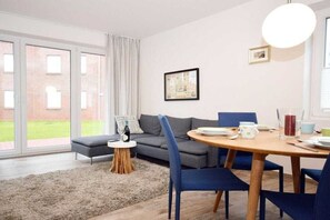 A modern living room with a sofa, a round wooden dining table, and chairs. There is a small side table with a bottle and a glass. The room has a large window with a view of a building outside. - Holiday apartment with 2 bedrooms (Norden-Norddeich)