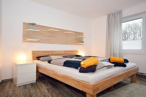 A modern bedroom with a wooden bed, bedside tables, a wall-mounted light fixture, and a window with curtains. - Holiday apartment with 2 bedrooms (Norden-Norddeich)