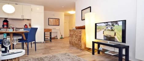 Een moderne woonkamer met een televisie, een eethoek met een tafel en stoelen, en een keuken met kasten en apparatuur.