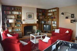 Een woonkamer met rode meubels, een open haard, een televisie en boekenkasten vol boeken.