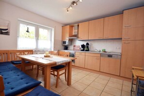A kitchen with wooden cabinets, a dining table with blue cushioned benches, and a window with curtains. - Holiday home with 2 bedrooms (Norddeich)