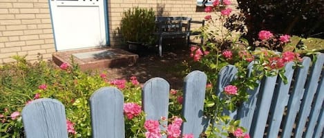 A brick building with a white door, a wooden bench, and a wooden fence with pink flowers.