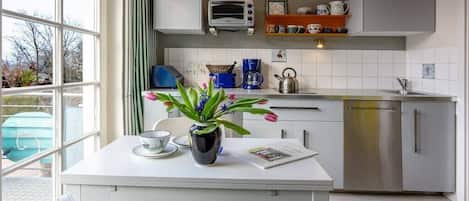 Une cuisine moderne avec un plan de travail blanc, une bouilloire, une machine à café et un grille-pain. Une fenêtre offre une vue sur l’extérieur.