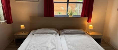 A double bed with a headboard, two bedside tables with lamps, a window with red curtains, and a framed picture on the wall.