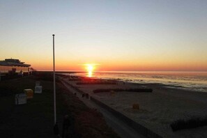 Um pôr do sol sobre uma praia, com uma passarela que leva à água, um poste alto e algumas construções à esquerda.