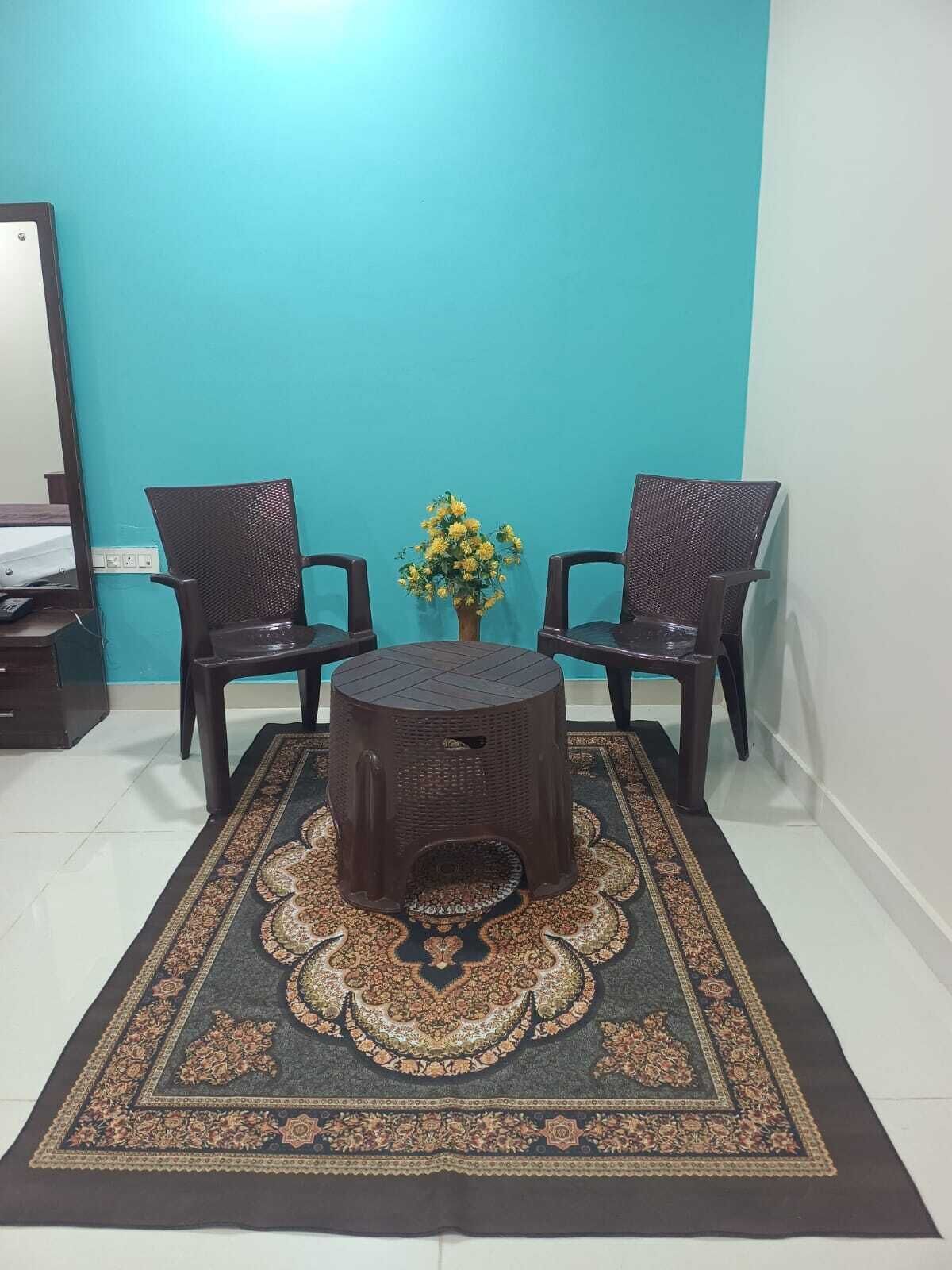 A room with two chairs, a small stool, a vase with yellow flowers, and a patterned rug on a tiled floor.