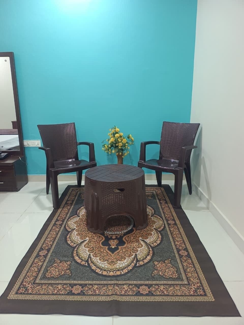 A room with a blue wall, two wicker chairs, a round table, and a patterned rug.