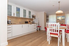 Eine moderne Küche mit weißen Schränken, einer Mikrowelle und einer Kaffeemaschine. Es gibt eine Essecke mit einem Holztisch und weißen Stühlen mit roten Kissen.