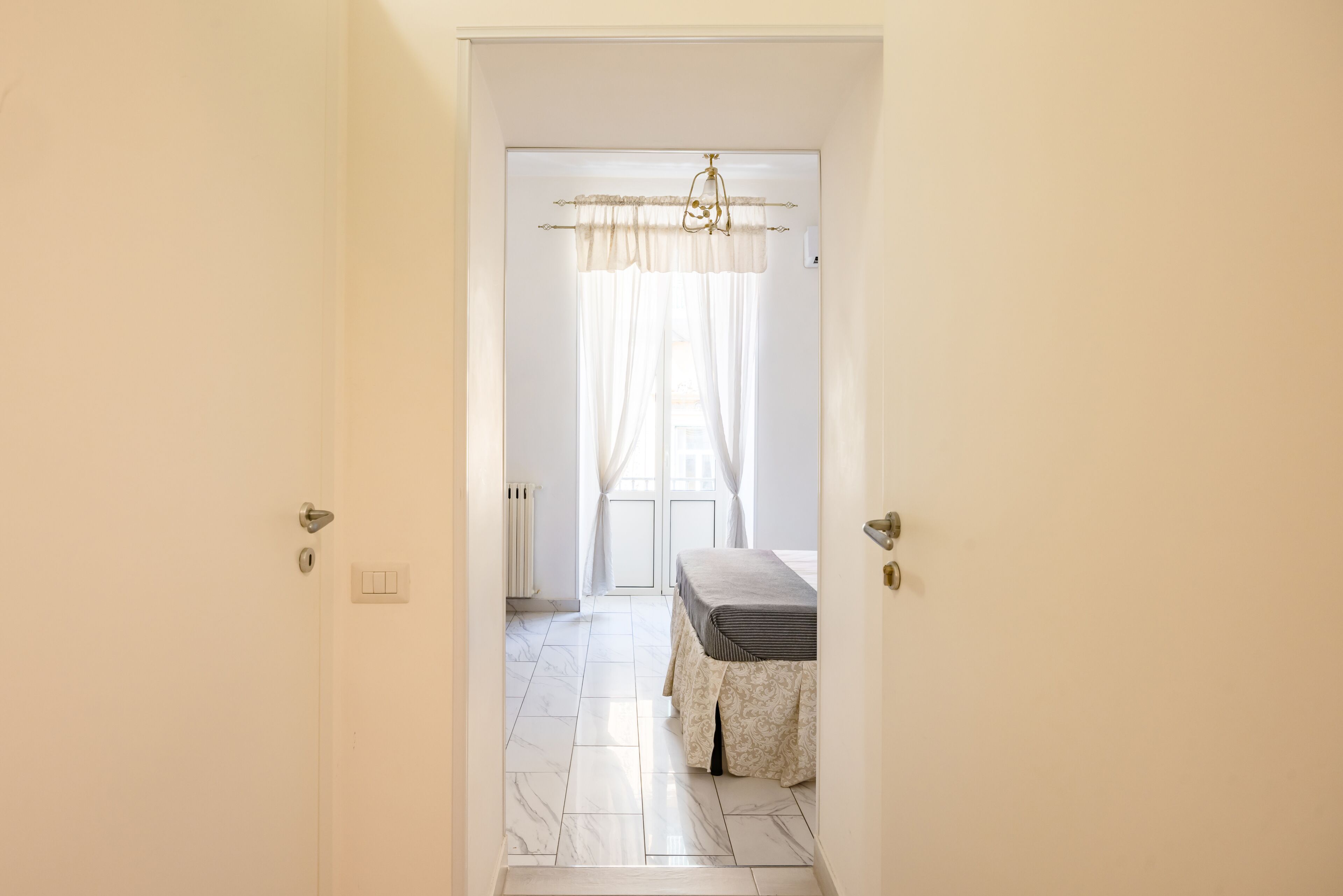 Un pasillo que lleva a un dormitorio con una cama, un radiador y una ventana con cortinas.