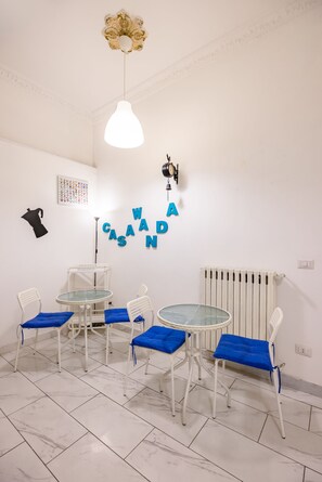 Een kamer met een wit plafond, een hanglamp, een wandklok en een radiator. Er staan twee ronde tafels met blauwe kussens en witte stoelen.