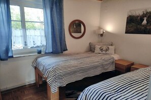 Ein Schlafzimmer mit zwei Betten, einem fensterförmigen Ausblick mit Vorhängen, einer Wandlampe und einem gerahmten Bild.