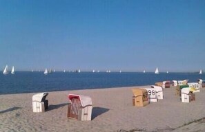 Ein Strand mit mehreren kleinen, nummerierten Strandkörben und Segelbooten in der Ferne.