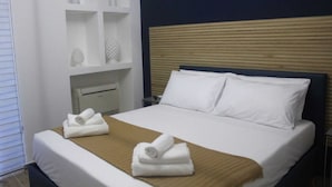 Habitación de hotel con cama, almohadas blancas, cabecera de madera, mesita de noche y estante con objetos decorativos.