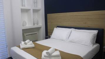 A hotel room with a bed, white pillows, a wooden headboard, a bedside table, and a shelf with decorative items.