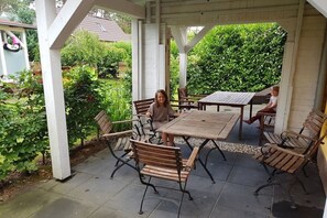 Een terras buiten met een houten tafel en banken, omgeven door groen.