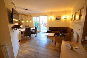 Ein Wohnzimmer mit einer Couch, einem Esstisch und Stühlen. Ein Fernseher ist an der Wand befestigt.