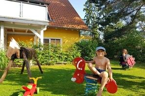 Criança brincando com um cavalo de brinquedo e um balanço em um jardim, com uma casa amarela ao fundo.