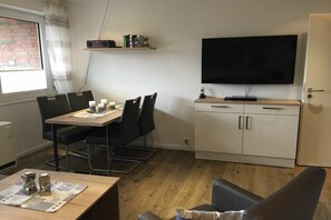 A room with a dining table, chairs, a flat-screen TV mounted on the wall, a cabinet with drawers, and a window with curtains. - Holiday apartment with 1 bedroom (Norden)