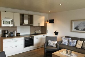 A modern kitchen with white cabinetry, a built-in oven, and a microwave. There is a grey sofa with patterned cushions and a coffee table with a newspaper. - Holiday apartment with 1 bedroom (Norden)