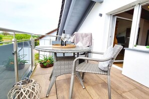 Een balkon met een glazen tafel, rieten stoelen, een wijnfles en een glas.
