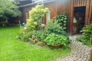 Ein Garten mit verschiedenen Pflanzen und Blumen, ein hölzernes Gebäude und ein Pflasterweg.