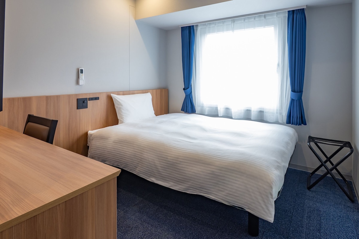 A hotel room with a bed, a wooden desk, a chair, a small table, and a window with curtains.