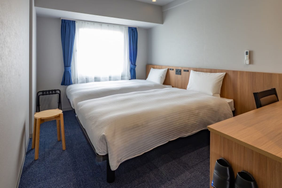 A hotel room with two beds, a desk, a chair, a small table, and a window with curtains.
