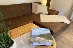 Ein braunes Ledersofa mit einer beige farbenen Decke und Kissen, ein Couchtisch aus Holz mit einem Buch mit dem Titel 'Exquisite Ferienwohnungen an der Ostsee' und eine Topfpflanze.