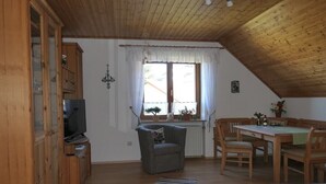 Ein gemütliches Zimmer mit Holzbalkendecke, einem Fernsehgerät auf einem Schrank, einem Sofa, einem Esstisch mit Stühlen und einem Fenster mit Vorhängen.