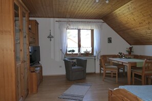 Una habitación acogedora con techo de madera, un televisor sobre un mueble bajo, un sofá, una mesa de comedor con sillas y una ventana con cortinas.