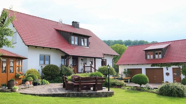 Uma casa de dois andares com telhado de telhas vermelhas, varanda frontal com um banco e um jardim bem cuidado.