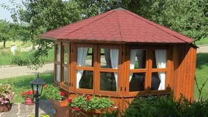 Um gazebo de madeira com telhado vermelho, cercado por vegetação exuberante e plantas em vasos.
