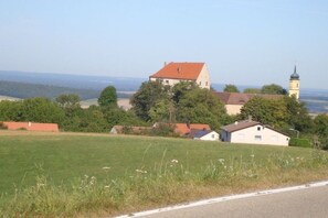 Paisaje rural con un edificio destacado de techo rojo, una iglesia con campanario y una carretera en primer plano.