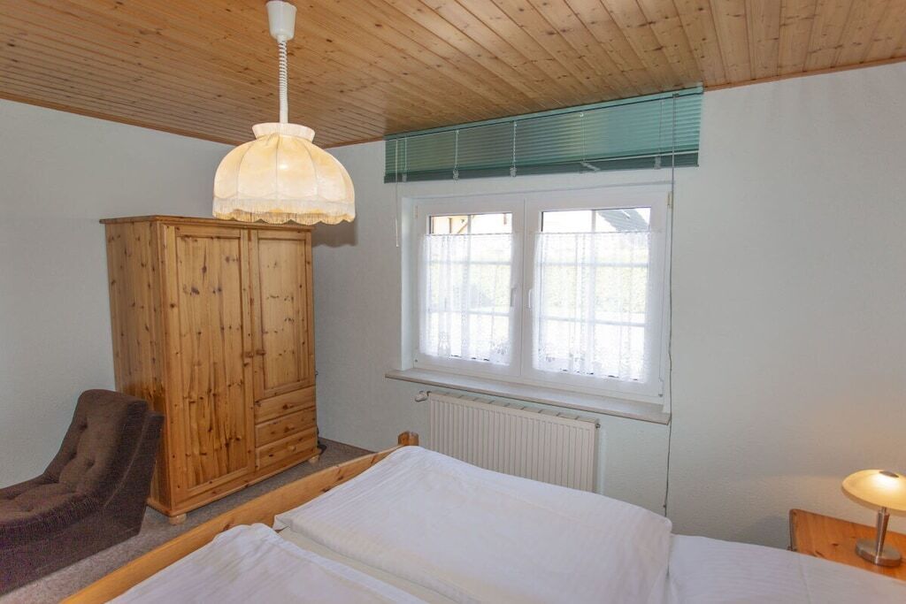 Een slaapkamer met een houten garderobe, een bed, een stoel, een raam met jaloezieën en een hanglamp.